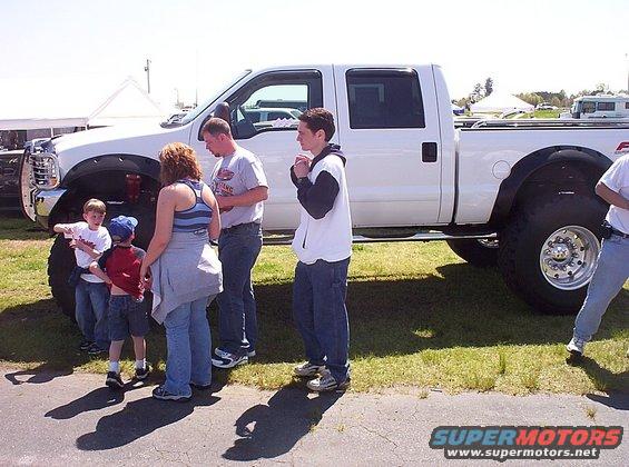 People stand around a lifted white pickup truck with large tires