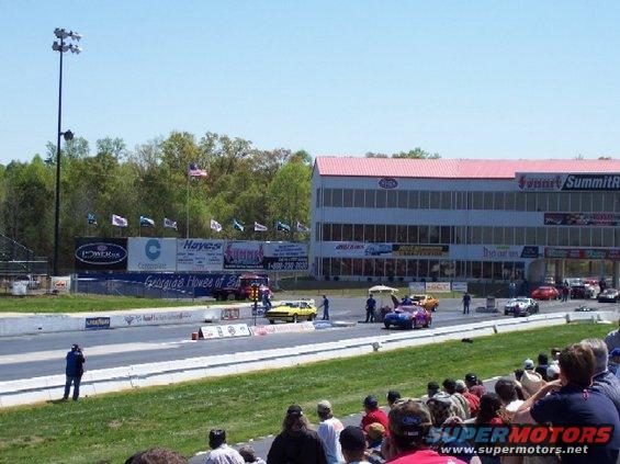 Drag strip with multiple cars, spectators, and a grandstand building
