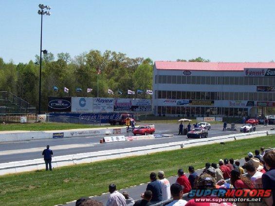 Drag strip with multiple cars racing, grandstands, and a large building in the background