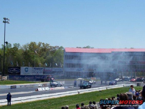 Cars racing on a drag strip with a grandstand and building in the background