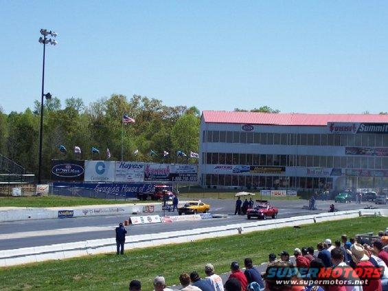 Drag strip with multiple cars, grandstands, and a multi-story building. Watermark: SUPER MOTORS www.supermotors.net