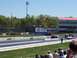 1978 Ford F-250 - Cars compete on a drag strip under a clear blue sky, with spectators in the foreground and a large building in the background.