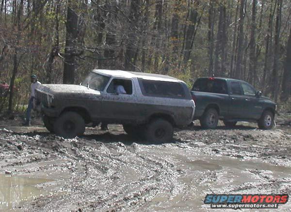 Mud-covered 1980s Ford Bronco with a white top, stuck in mud next to a dark green pickup truck