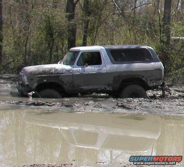 Mud-covered 1978 Ford Bronco stuck in mud, side view. Water reflection visible