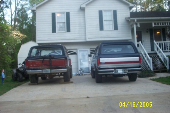 two Ford Broncos parked in front of a house, one red and black, one blue and silver