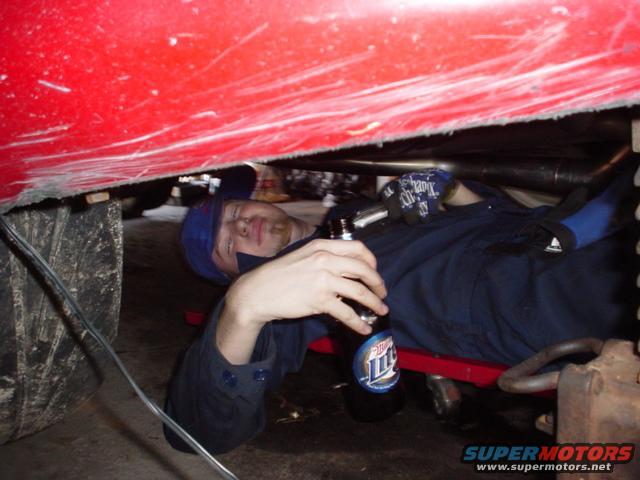 Man in blue overalls and cap lying on creeper under red car, holding a bottle of Miller Lite