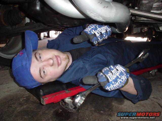 Guy in blue jumpsuit and red cap working on car underside with wrenches