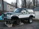 1994 Ford Bronco - A two-tone Ford Bronco is elevated on jack stands in a driveway, showing its front end.