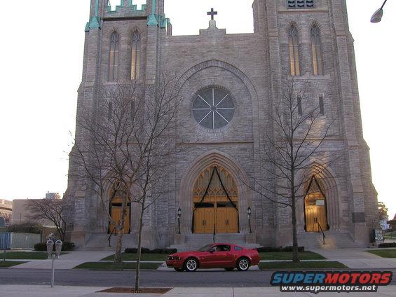 Red 2005 Ford Mustang parked on street in front of a large stone church with yellow doors