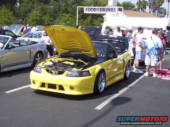 Yellow convertible Mustang with hood open, parked next to a silver convertible. People milling around