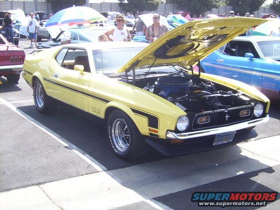 Yellow 1971 Ford Mustang Mach 1 with hood open, showing engine. Black stripe on side. Blue car in background