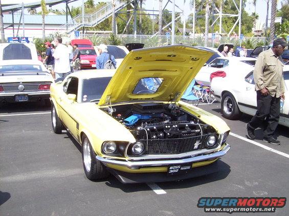 Yellow 1969 Ford Mustang Boss 302 with hood open, showing engine. Other cars and people in background