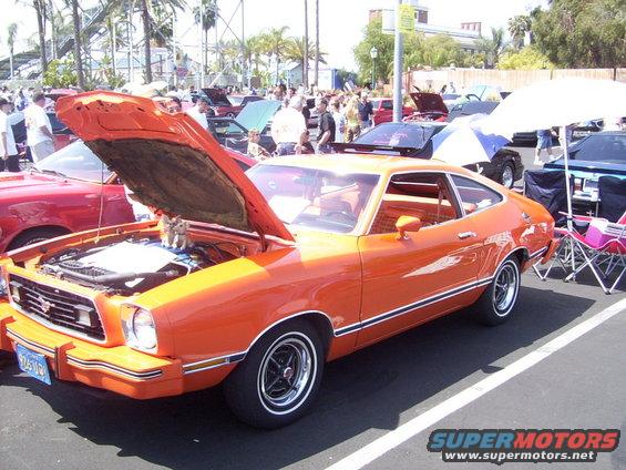 orange coupe with hood open, showing engine. Other cars and people in background at an outdoor event