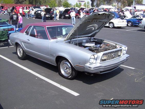 Silver 1975 Ford Mustang coupe with maroon roof, hood open showing engine, parked at an outdoor car show