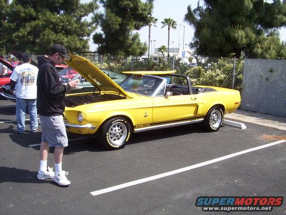 Yellow 1968 Ford Mustang convertible with hood open, GT350 stripe. Two people standing nearby
