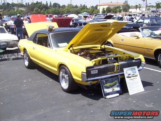 Yellow classic car with black roof, hood open showing engine. Display board in front