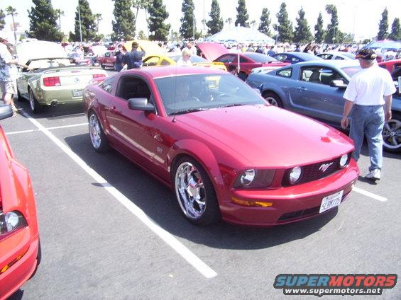 Red Mustang parked at a car show. Other cars and people are visible in the background