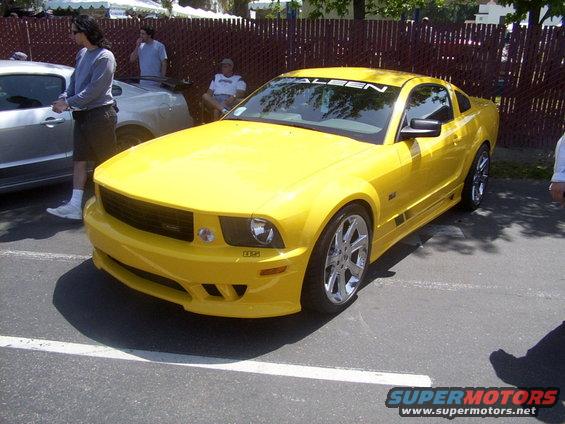Yellow Saleen Mustang coupe parked on asphalt, front 3/4 view. Aftermarket body kit and wheels are visible