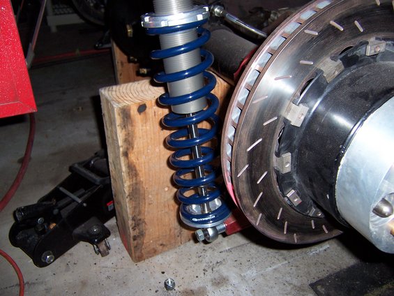 Close-up of a blue coilover shock absorber and a slotted brake rotor, supported by wooden blocks and a floor jack