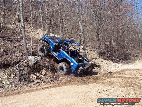 Blue buggy with roll cage going up a steep, wooded hill. One front wheel is off the ground