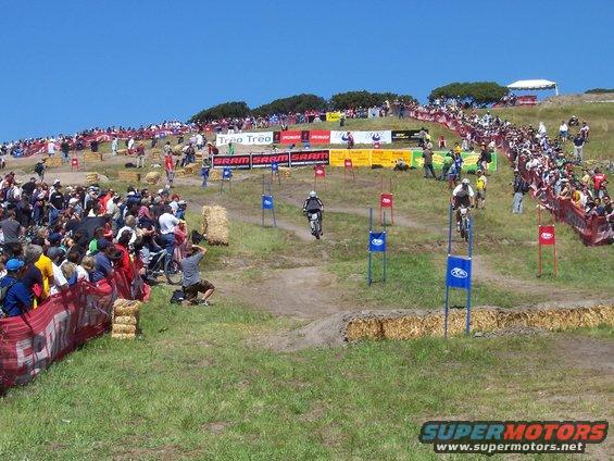 Crowd watching a downhill mountain bike race on a grassy hill with banners and hay bales