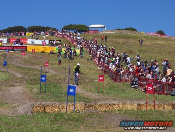 Cyclist in yellow jersey on a dirt track, with hay bales and spectators lining the course
