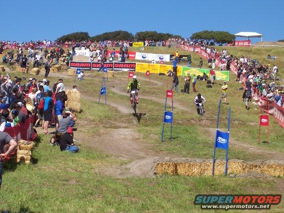 Mountain bikers race downhill on a grassy course with spectators lining the hillsides