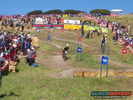 People watching a mountain bike race on a grassy hill with hay bales and banners