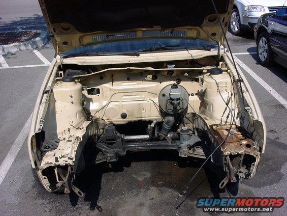 Tan car engine bay with hood open, showing disassembled front end and engine components. Rust is visible
