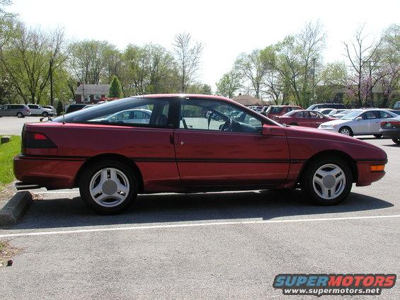 Red coupe parked in a lot, side view. The car has a sloping roofline and distinctive wheels