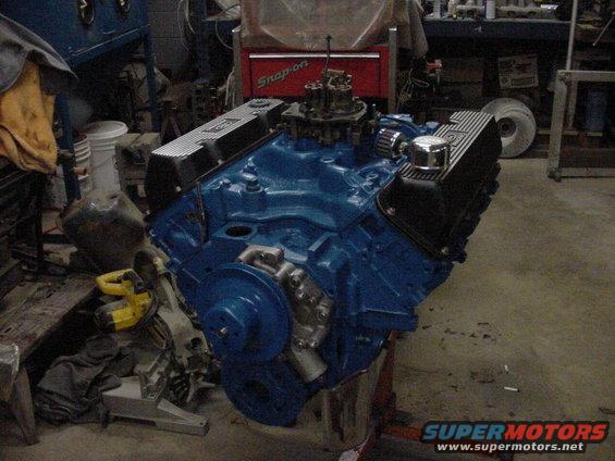 Blue V8 engine with chrome valve covers and carburetor on an engine stand in a workshop