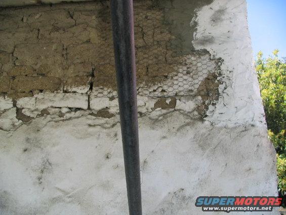 Damaged stucco wall with exposed brick and wire mesh, a metal pipe runs vertically