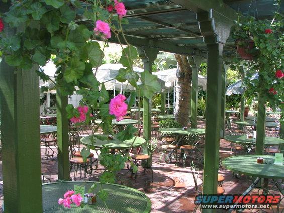 Outdoor cafe seating area with green tables and chairs, pink flowers, and a Supermotors.net watermark