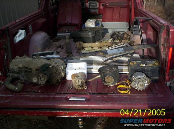 Various car parts including a transmission, starter, alternator, and air filter are laid out in the bed of a red pickup truck