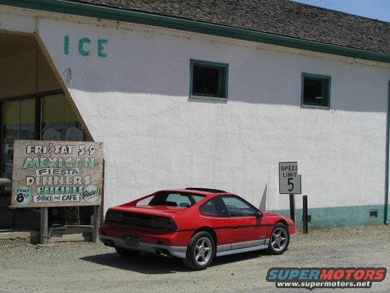 Red Pontiac Fiero parked outside a building with 'ICE' painted on the side. Sign advertises Mexican dinners