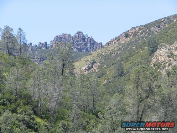Rocky hills with scattered trees under a clear blue sky. Supermotors logo in lower right