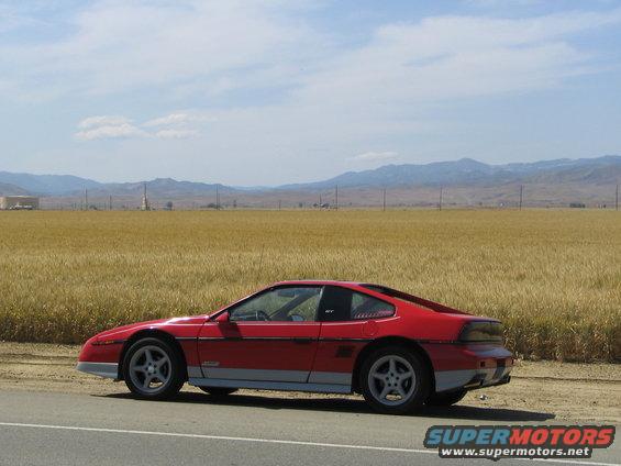 Red 1986 Pontiac Fiero parked on the side of a road with a field of wheat and mountains in the background