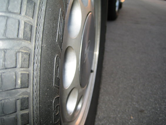 Close-up of a silver wheel with circular cutouts and a tire on a paved surface. Another vehicle is visible in the background