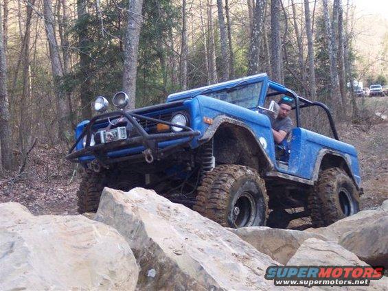 Blue SUV with off-road tires navigating large rocks on a wooded trail. Driver visible