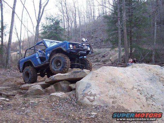 Blue 1974 Ford Bronco with roll cage and off-road tires climbing over large rocks