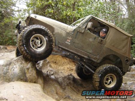 Mud-covered Jeep Wrangler climbing a rock obstacle, driver visible