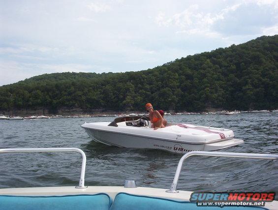 Two boats on a lake, one with two people, wooded hills in background. Supermotors.net logo visible