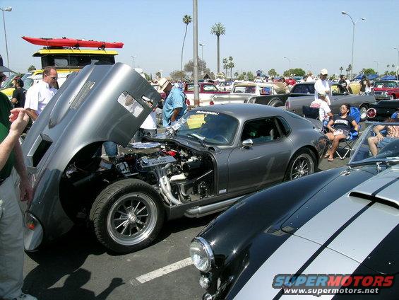 Gray coupe with hood open, engine exposed. Another car with black and silver stripes in foreground