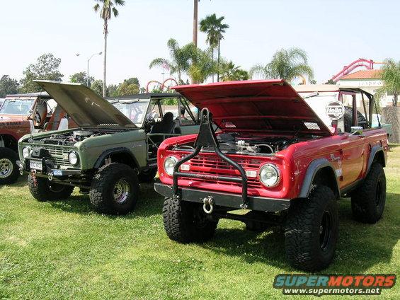 Two Ford Broncos, one red and one green, parked on grass with hoods open. A third Bronco is partially visible to the left
