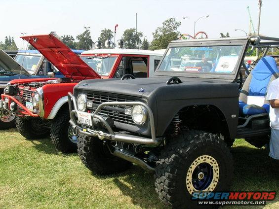Modified black Ford Bronco with off-road tires and roll cage, parked next to a red Ford Bronco with hood open. License plate reads LHTL 691