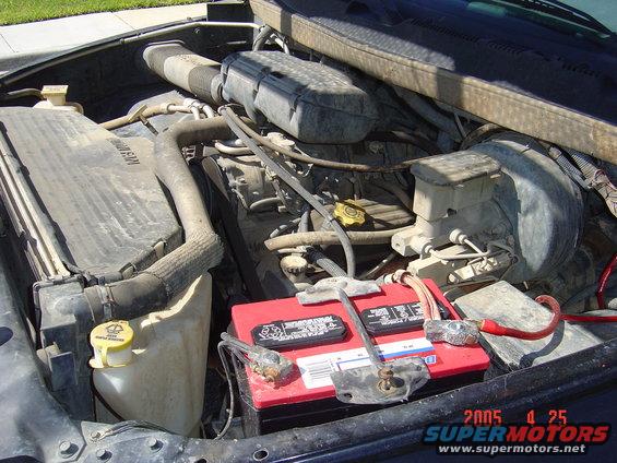 Engine bay with a red battery, hoses, and engine parts. Date stamp says 2005