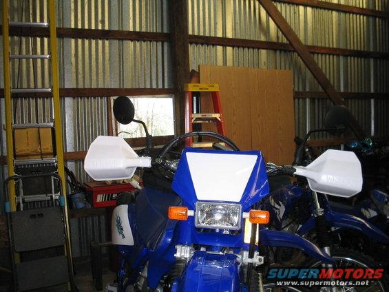 Front view of a blue dirt bike with white handguards and a white windscreen. Another blue motorcycle is visible behind it