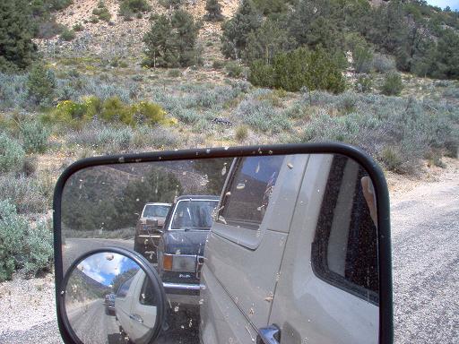 Side view mirror reflection shows two vehicles behind the camera vehicle on a dirt road