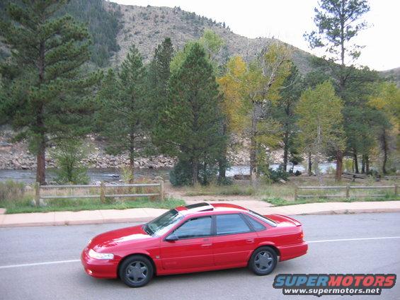 red sedan parked on roadside with trees and river in background