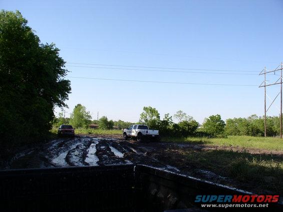 Two trucks, one white and one red, driving through a muddy field under a clear blue sky